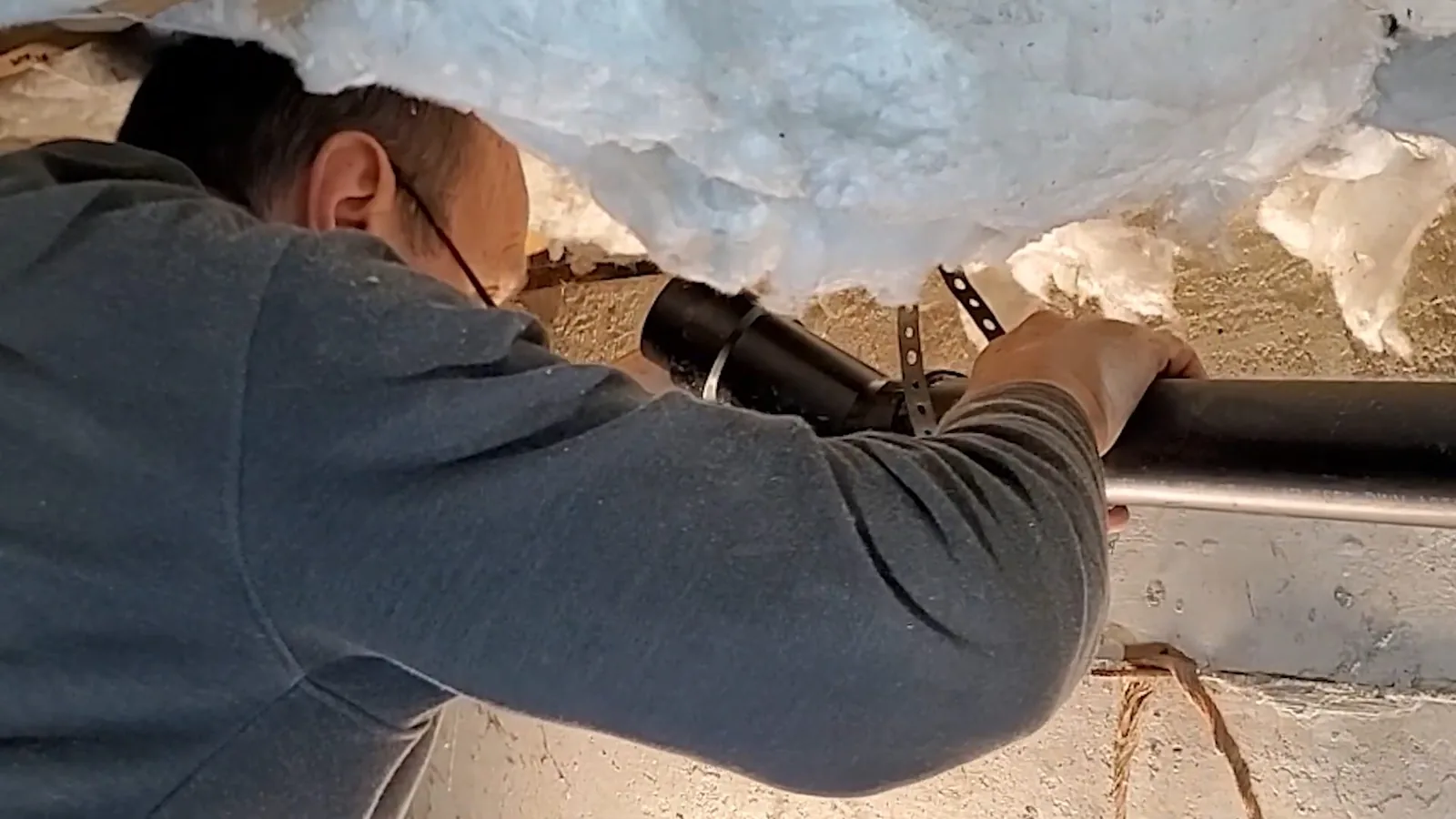 Technician with his arm through blown insulation reaching a black drain pipe in a low space