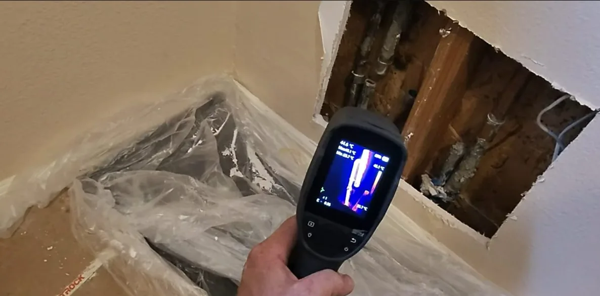 Hand holding a thermal camera toward an opened wall cavity of pipes, with a cool streak on its screen