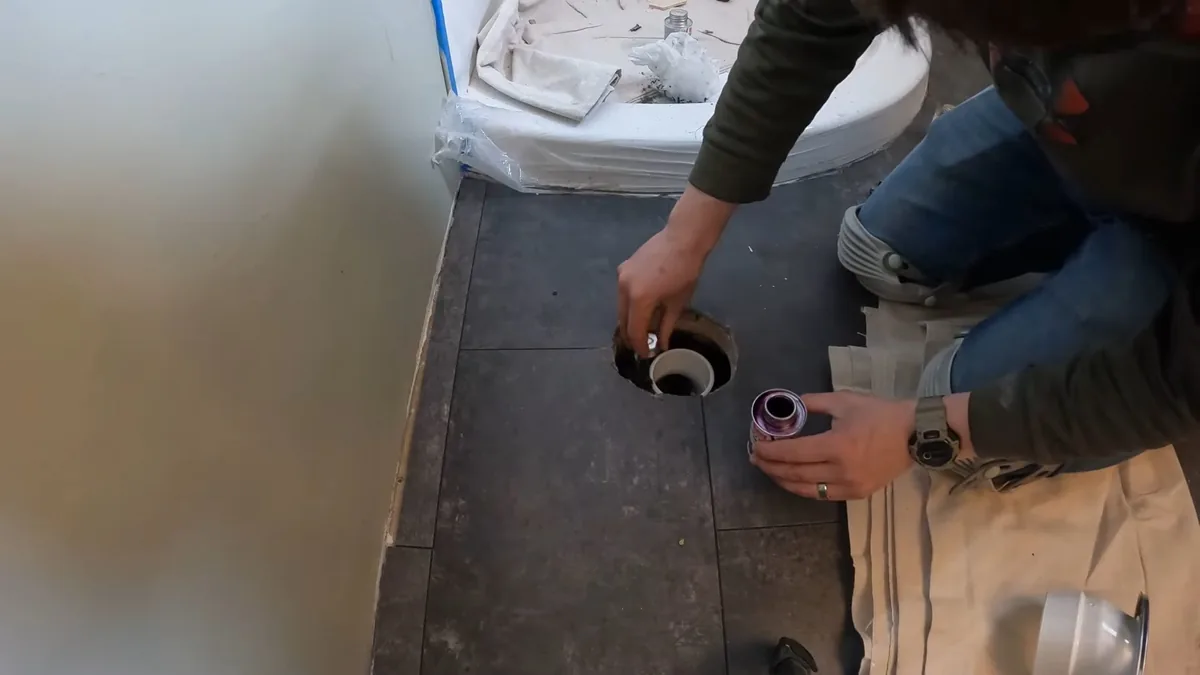 Person kneeling on a tiled floor beside a toilet drain flange with a can of purple primer next to it