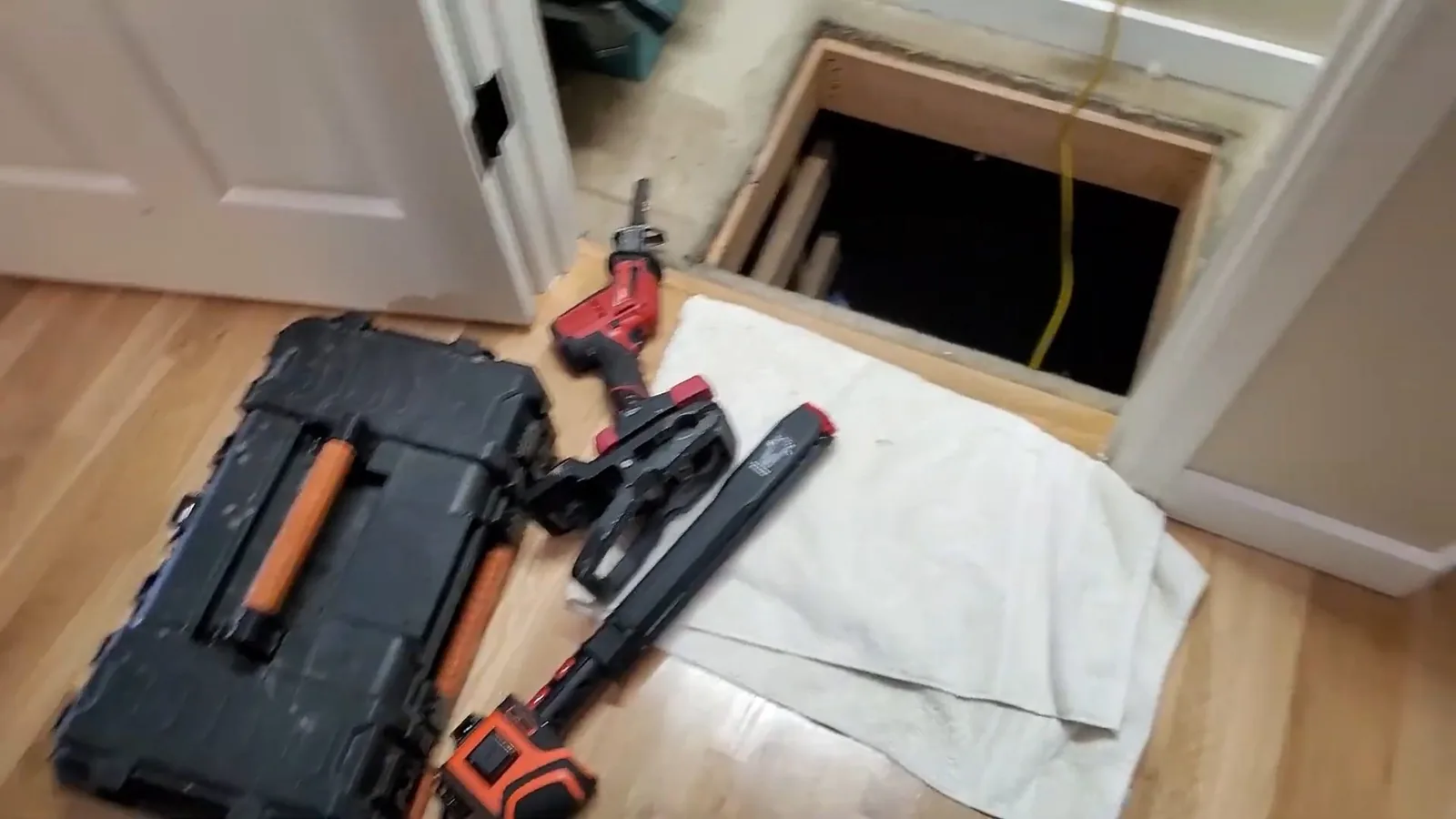 Black toolbox, cordless saw and wrench laid on a towel on a wood floor beside an opened floor access hole