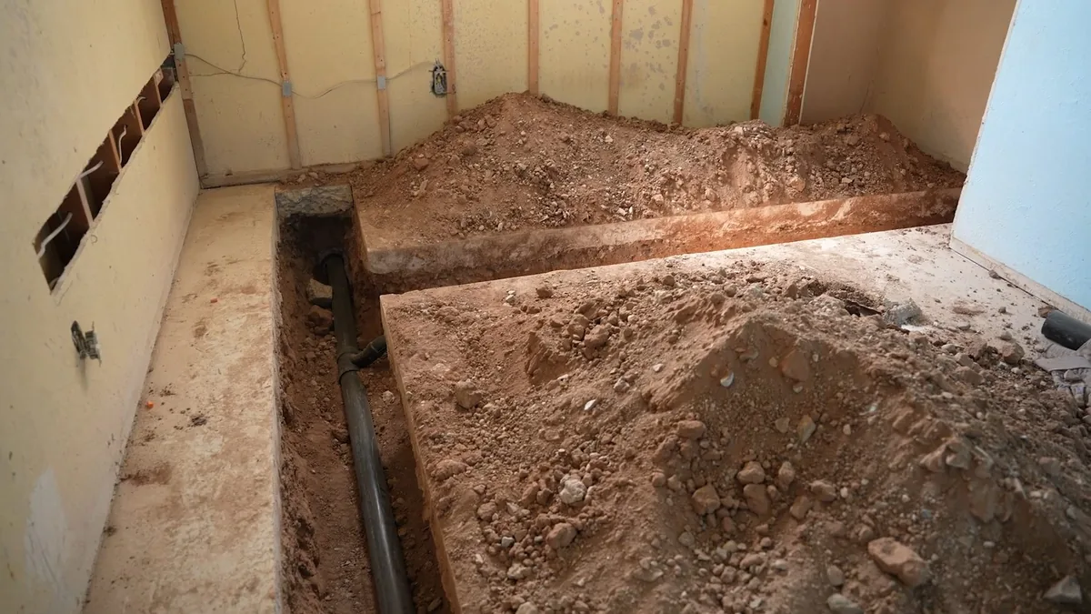 Trench cut through a floor slab with a dark pipe laid along the bottom and piles of soil on either side