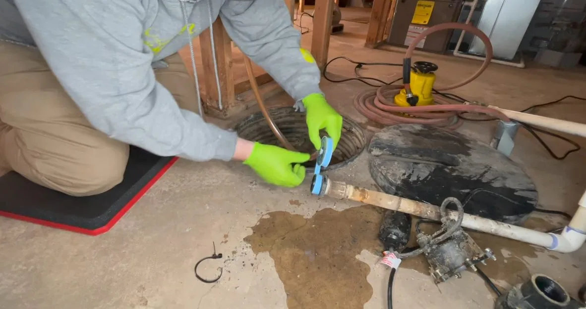 Person in green gloves working at an open sump pit with brown water pooled on the basement floor