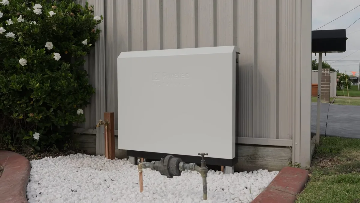 White outdoor equipment cabinet mounted on a siding wall above white gravel beside a flowering shrub