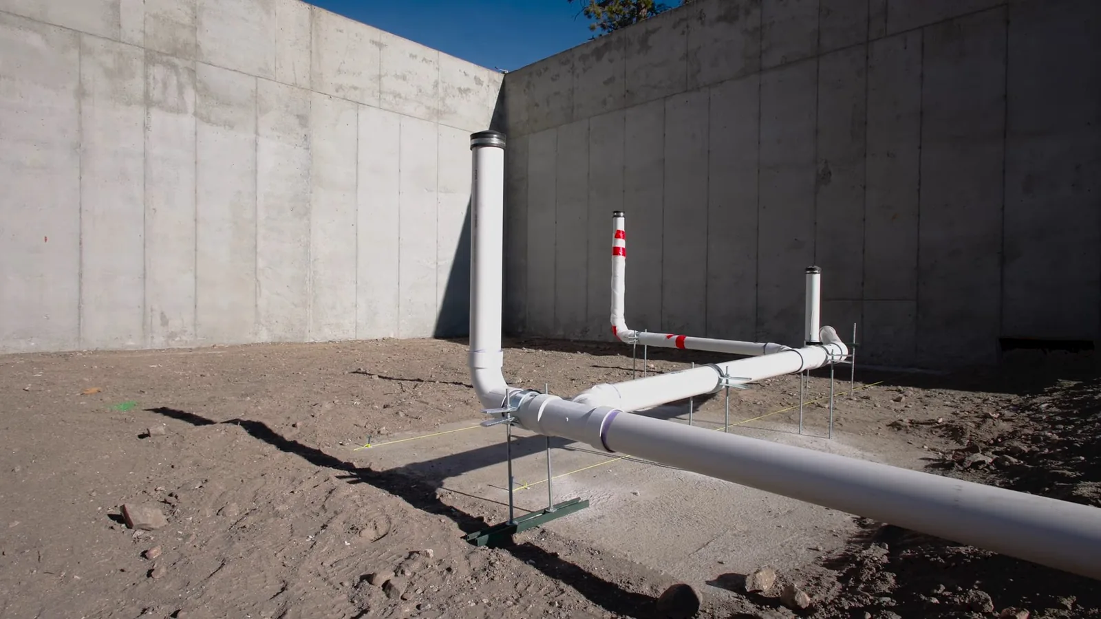 White PVC drain and vent stubbed up from bare graded ground beside a tall concrete wall