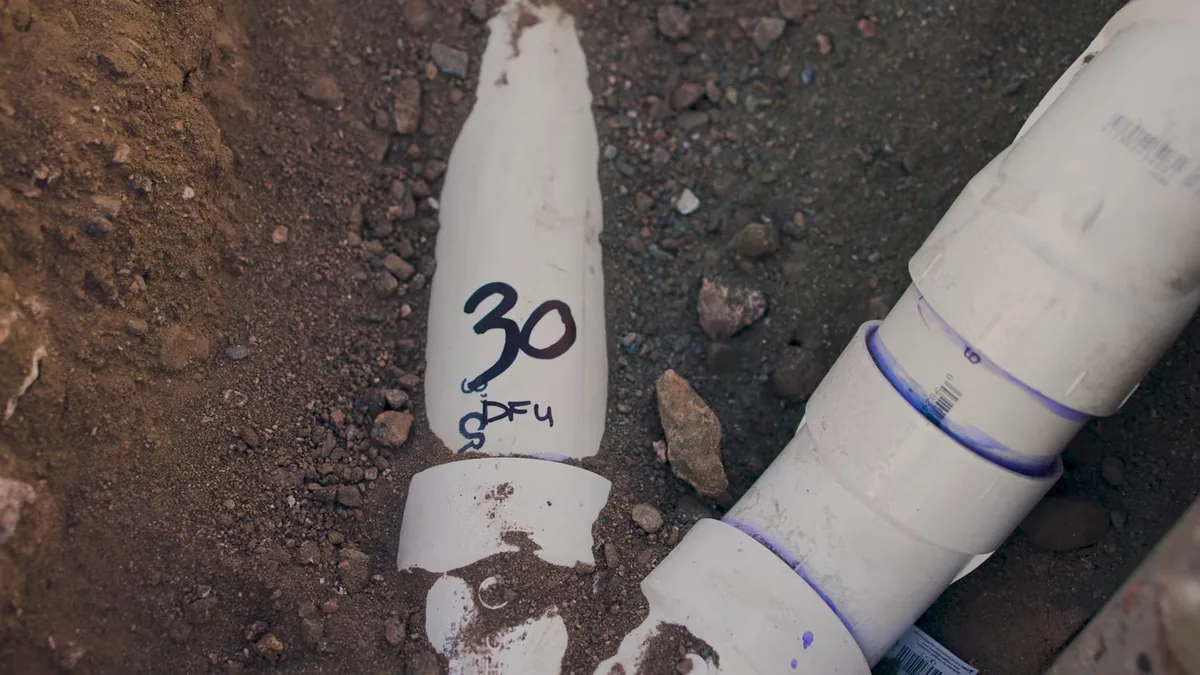 White PVC pipe fittings with purple primer joints and a marked cut section lying in a soil trench