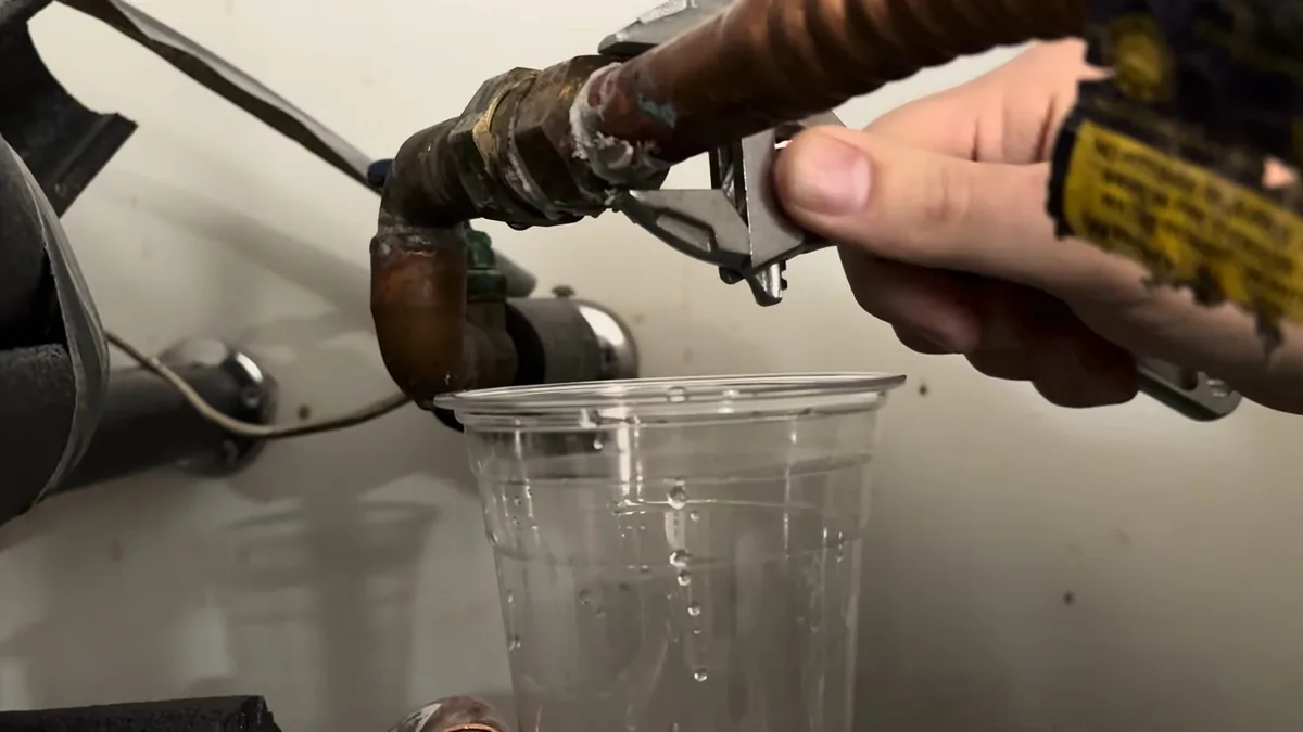 Hand with an adjustable wrench opening a drain fitting above a clear plastic cup beside a water heater
